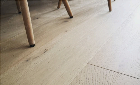 Close-up of light-colored wooden floorboards with visible wood grain and knots. The legs of a piece of furniture with wooden legs and black feet are partially visible at the top of the image.