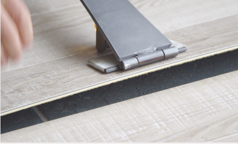 A close-up of flooring installation; a metal tool presses down on a plank of light wood laminate as a hand works nearby, with underlayment visible beneath the floorboards.
