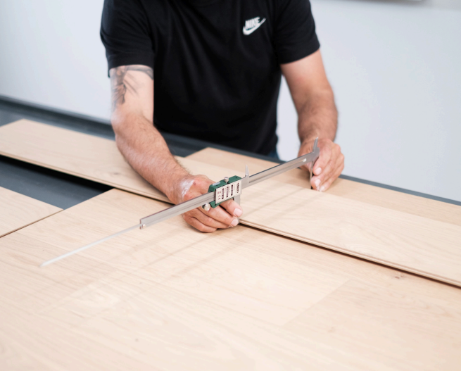 A person in a black shirt uses a digital caliper to measure the width of a light wooden plank placed on a table.
